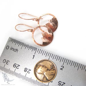 May include: A pair of rose gold earrings with a hammered metal design. Each earring features a circle with a mountain silhouette cut out. The earrings are shown next to a ruler and a US penny for size reference.