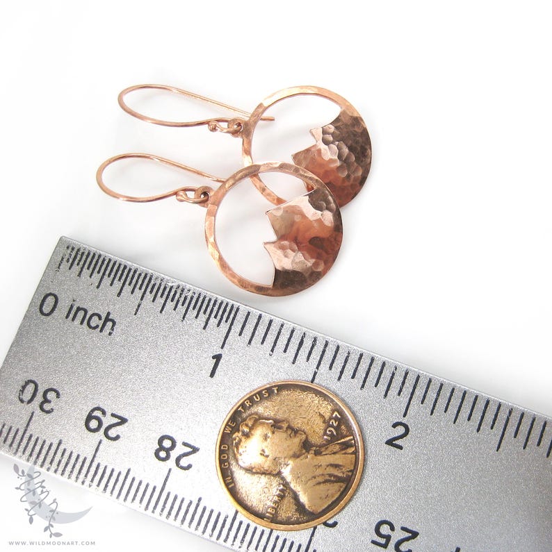 May include: A pair of rose gold earrings with a hammered metal design. Each earring features a circle with a mountain silhouette cut out. The earrings are shown next to a ruler and a US penny for size reference.