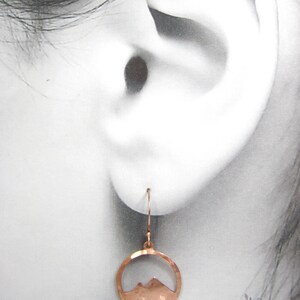 May include: Copper earrings with a hammered finish and a mountain silhouette cutout. The earrings are hanging from a person's ear.