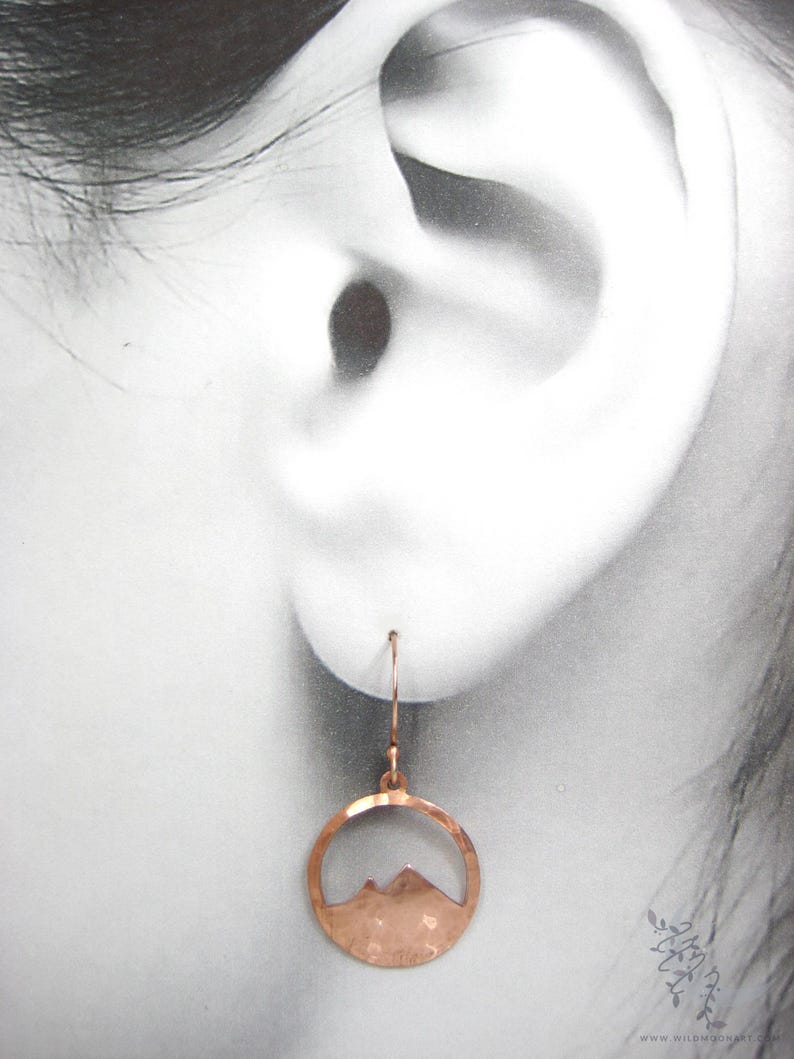 May include: Copper earrings with a hammered finish and a mountain silhouette cutout. The earrings are hanging from a person's ear.
