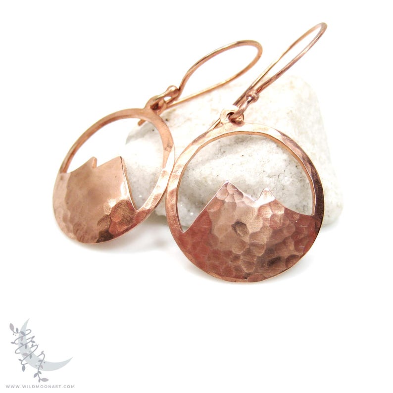 May include: A pair of copper earrings with a hammered finish. Each earring features a circular design with a mountain silhouette cut out. The earrings are suspended from a simple wire loop.