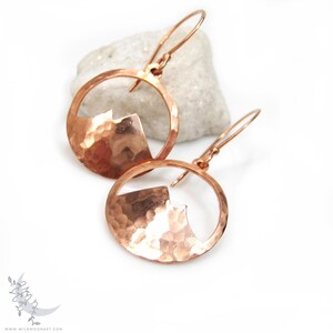 May include: A pair of copper earrings with a hammered, circular design. Each earring features a cut-out shape resembling a mountain peak.