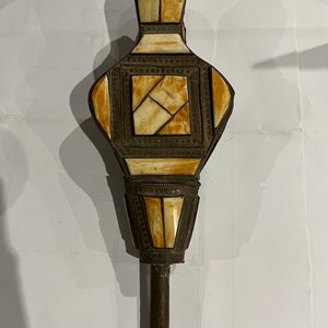 Op de afbeelding: Een decoratief, versierd, ivoor-kleurig, metalen, handgehouden object met een lange, dunne handgreep. Het object is traanvormig en heeft ingewikkelde geometrische patronen.