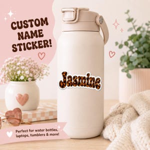 May include: A beige insulated water bottle featuring a custom name sticker with the name Jasmine written in a wavy font with a brown and black tortoiseshell pattern. The sticker has a white border and is applied to the center of the bottle.