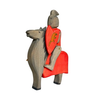 May include: A wooden toy knight riding a gray horse. The knight is wearing red armor and holding a shield with a dragon design.
