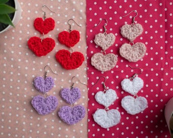 Pode incluir: Quatro pares de brincos de crochê feitos à mão em formato de coração nas cores vermelho, rosa claro, lavanda e branco. Cada brinco apresenta dois motivos em formato de coração empilhados, conectados por ganchos de metal prateado.