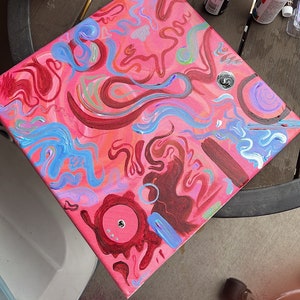 May include: A pink canvas painting with abstract swirls and lines in shades of blue, red, and green. The painting is on a table with paint bottles and brushes.