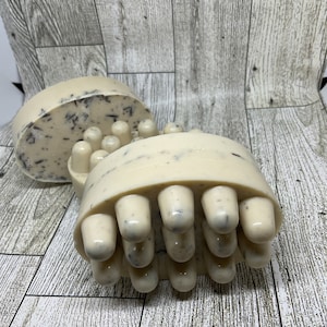 May include: Two white and gray exfoliating soap bars with raised bumps on one side. The soap bars are on a light wood background.