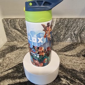 May include: A white water bottle featuring a colorful design with the word "ROBLOX" and various cartoon characters. The bottle has a blue handle and lid, with a green section near the top. It is sitting on a white circular stand.