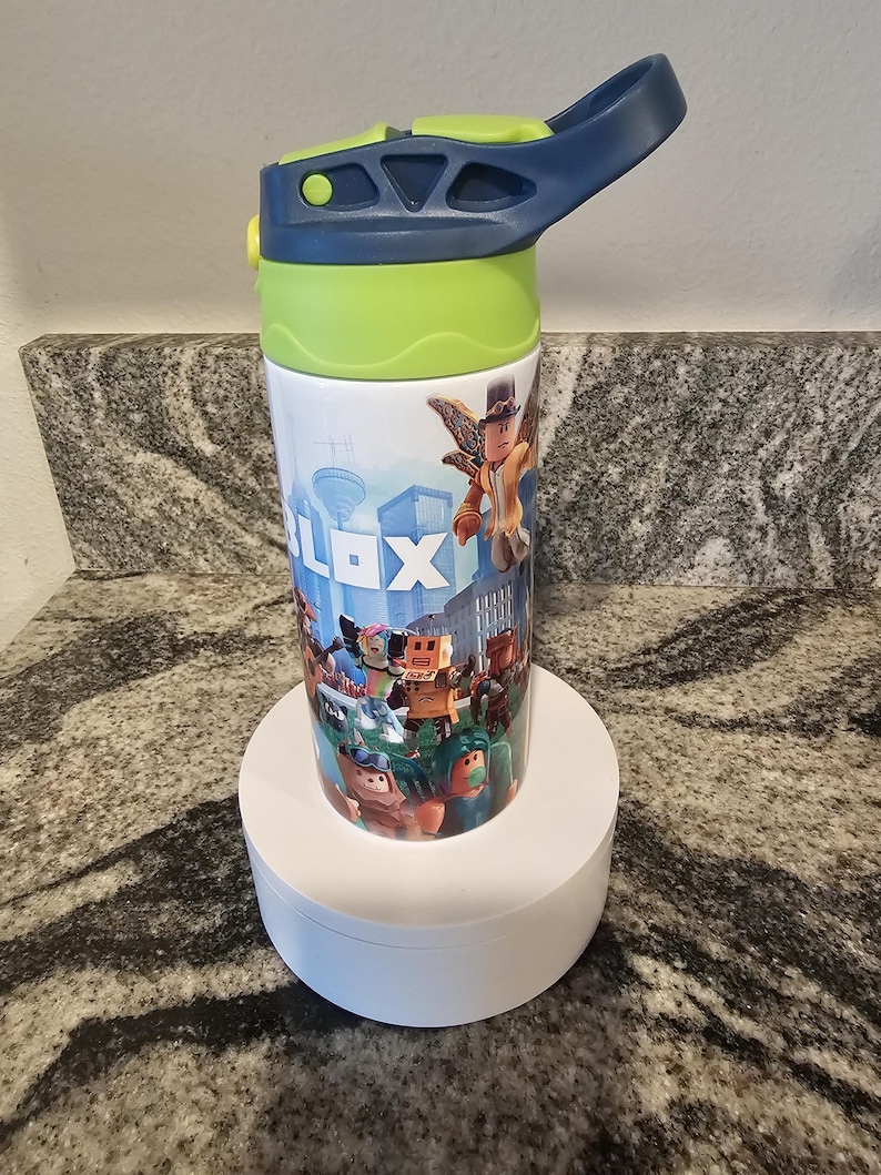 May include: A white water bottle featuring a colorful design with the word "ROBLOX" and various cartoon characters. The bottle has a blue handle and lid, with a green section near the top. It is sitting on a white circular stand.