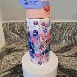 May include: A colorful water bottle featuring a cartoon character design. The bottle has a light blue, pink, and white lid with a push-button opening. The body of the bottle is decorated with cartoon characters, flowers, and glitter.