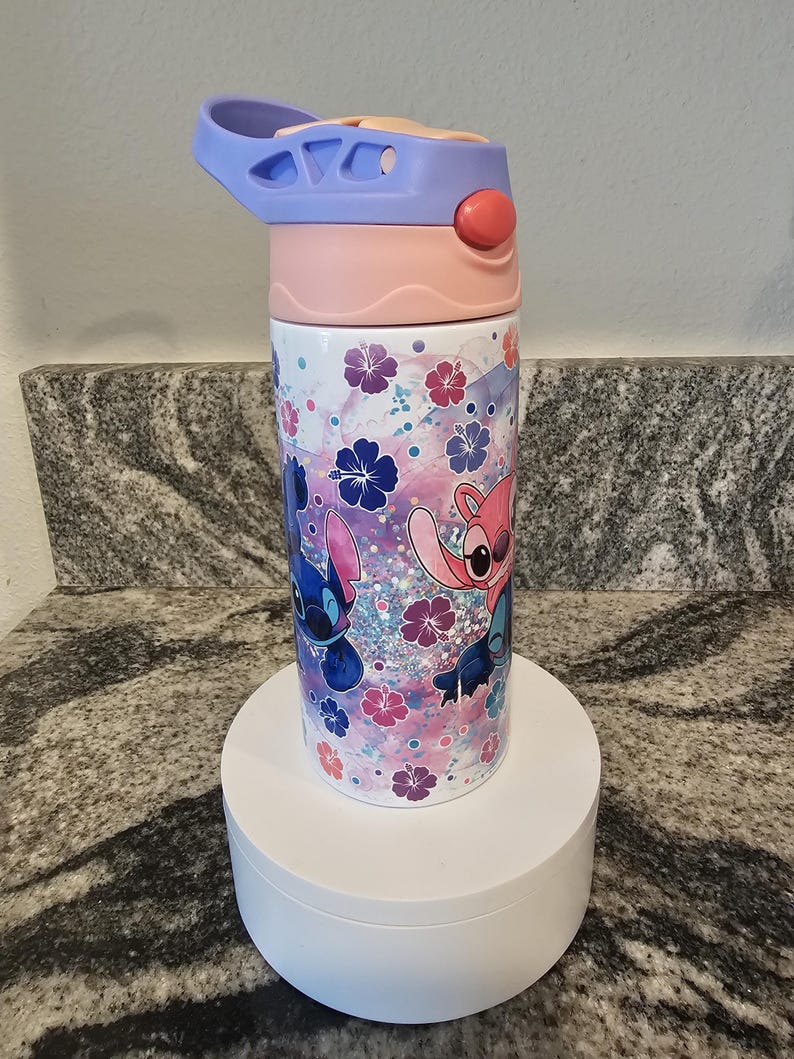 May include: A colorful water bottle featuring a cartoon character design. The bottle has a light blue, pink, and white lid with a push-button opening. The body of the bottle is decorated with cartoon characters, flowers, and glitter.