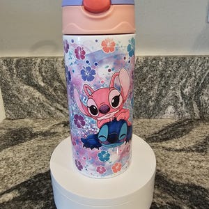 May include: A water bottle featuring a cartoon design of Stitch and Angel from Lilo & Stitch. The bottle has a pink, purple, and blue color scheme with a floral pattern. The lid is pink and purple.