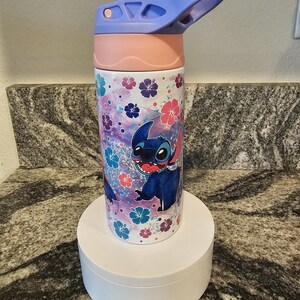 May include: A water bottle featuring a cartoon character, Stitch, with a floral pattern in pink, blue, and purple. The bottle has a pink lid and a blue flip-top. The bottle is cylindrical and sits on a white stand.