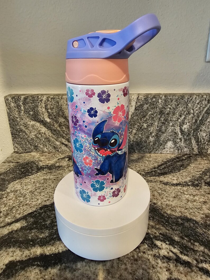 May include: A water bottle featuring a cartoon character, Stitch, with a floral pattern in pink, blue, and purple. The bottle has a pink lid and a blue flip-top. The bottle is cylindrical and sits on a white stand.