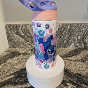 May include: A stainless steel water bottle featuring a cartoon character design. The bottle has a light purple handle and lid, a pink mid-section, and a white base. The design includes a blue cartoon character, flowers, and a glittery background.
