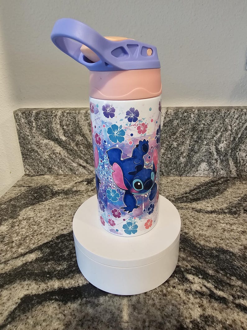 May include: A stainless steel water bottle featuring a cartoon character design. The bottle has a light purple handle and lid, a pink mid-section, and a white base. The design includes a blue cartoon character, flowers, and a glittery background.