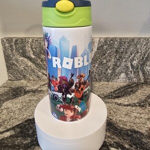 May include: A white water bottle featuring a colorful design with the word "ROBLOX" and various cartoon characters. The bottle has a blue and green lid with a yellow button. The bottle is cylindrical and sits on a white stand.