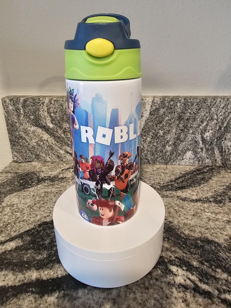 May include: A white water bottle featuring a colorful design with the word "ROBLOX" and various cartoon characters. The bottle has a blue and green lid with a yellow button. The bottle is cylindrical and sits on a white stand.