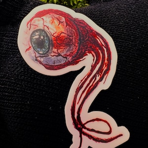 May include: A red and white sticker with a large, detailed eyeball with a blue iris and a red blood vessel pattern. The eyeball is attached to a long, thin, red and white tentacle that is curled at the end.