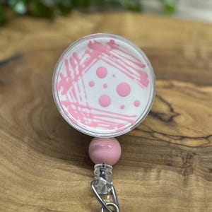May include: A pink and white petri dish design on a retractable badge reel. The badge reel has a pink ball and a silver clip. The petri dish design features pink streaks and dots.