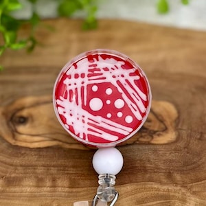 May include: A red and white badge reel featuring a petri dish design. The design includes white streaks and dots on a red background. The reel has a white, round top and a silver clip for attaching to clothing.