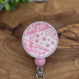 May include: Pink retractable badge holder with a clear plastic case and a pink and white abstract design. The case has a pink and white polka dot pattern.