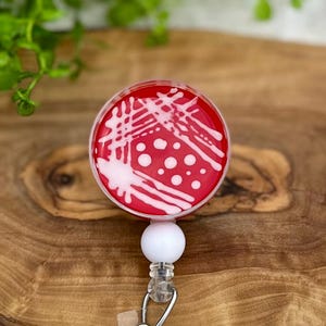 May include: A red and white badge reel with a petri dish design. The round badge features a red background with white patterns and dots, resembling bacterial growth. It has a white bead and a retractable cord with a metal clip.