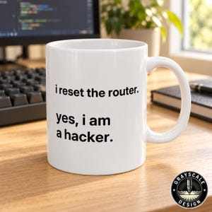 May include: A white ceramic mug featuring black text that reads, i reset the router. yes, i am a hacker. The mug has a standard cylindrical shape with a curved handle on the right side, presented against a blurred office desk background.