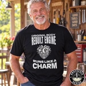 May include: A black short-sleeved t-shirt featuring white text that reads ORIGINAL BODY REBUILT ENGINE RUNS LIKE A CHARM. The design includes a graphic of a human heart constructed from mechanical gears and engine components centered on the chest.