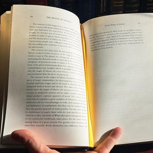 K&ouml;nnte beinhalten: Ein offenes Buch mit vergilbten Seiten, gehalten von den H&auml;nden einer Person. Der Text ist schwarz, mit dem Titel oben auf der rechten Seite sichtbar. Das Buch liegt auf einer dunklen Holzoberfl&auml;che, mit einem verschwommenen Hintergrund aus anderen B&uuml;chern.