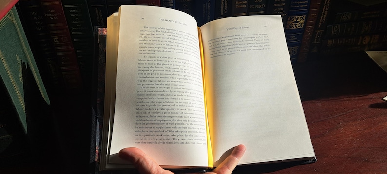 K&ouml;nnte beinhalten: Ein offenes Buch mit vergilbten Seiten, gehalten von den H&auml;nden einer Person. Der Text ist schwarz, mit dem Titel oben auf der rechten Seite sichtbar. Das Buch liegt auf einer dunklen Holzoberfl&auml;che, mit einem verschwommenen Hintergrund aus anderen B&uuml;chern.