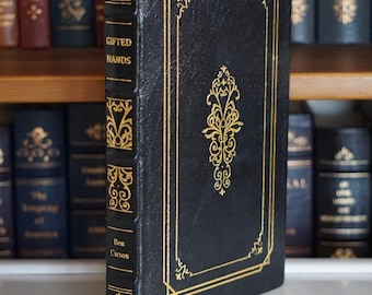 May include: A hardcover book titled "Gifted Hands" by Ben Carson. The book has a black cover with gold decorative accents and a gold bookmark. The book is standing upright on a white surface, with other books visible in the background.