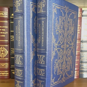 Puede incluir: Dos libros de tapa dura azules con letras doradas y dise&ntilde;os decorativos en las cubiertas. Los libros se titulan "Plutarch's Morals" y est&aacute;n en un estante de madera.