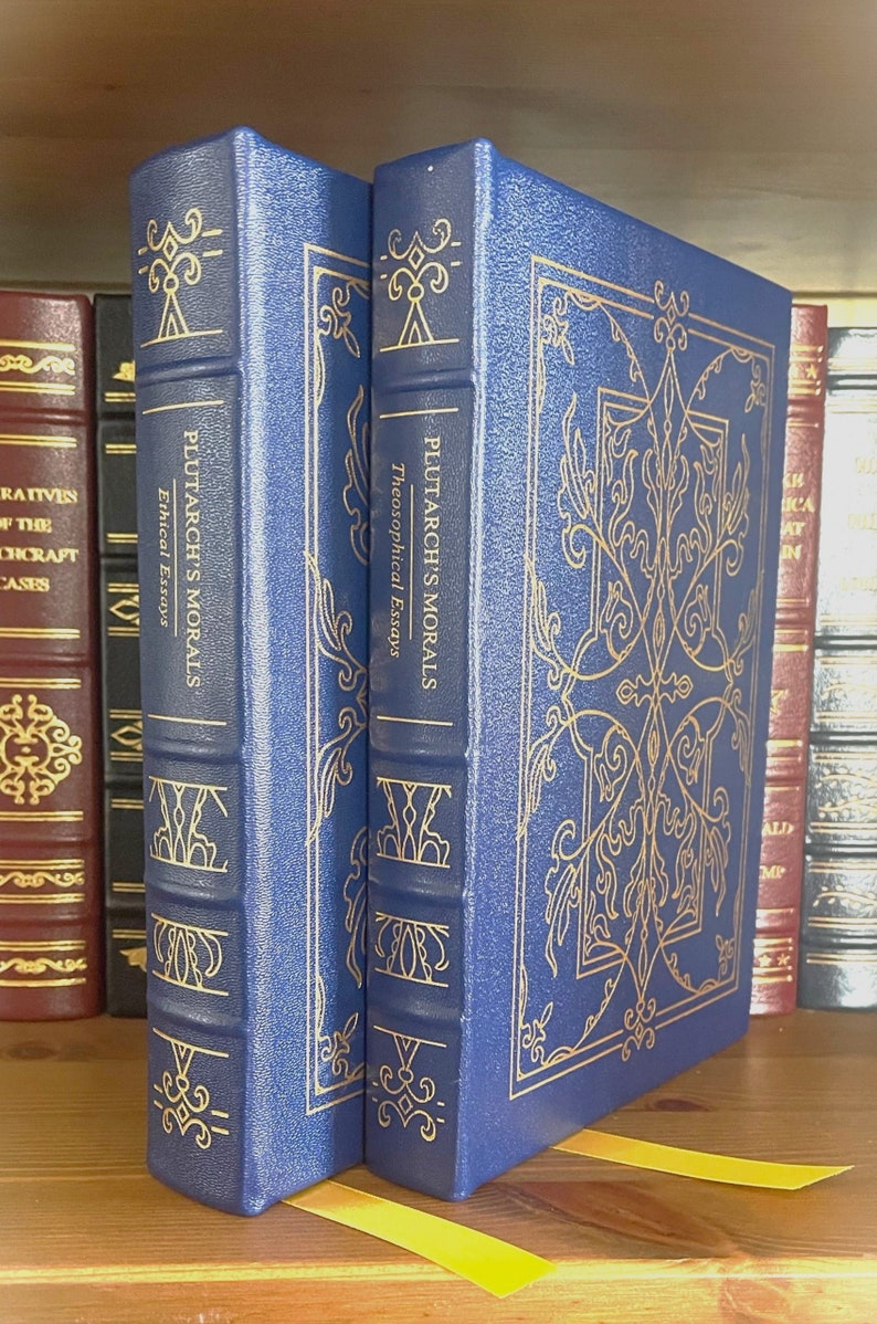 Puede incluir: Dos libros de tapa dura azules con letras doradas y dise&ntilde;os decorativos en las cubiertas. Los libros se titulan "Plutarch's Morals" y est&aacute;n en un estante de madera.