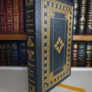 May include: A dark blue hardback book with gold accents and the title "On Classical Economics" by Thomas Sowell. The book is on a bookshelf.