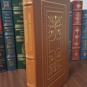 Puede incluir: Un libro encuadernado en cuero marr&oacute;n con letras doradas en el lomo. El t&iacute;tulo es "Greek Physiological Psychology" de W.H.S. Montague. El libro est&aacute; en un estante de madera con otros libros.