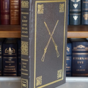 May include: A brown hardcover book with gold lettering and a gold design on the cover. The title of the book is "To Keep and Bear Arms" by Joyce Lee Malcolm. The book is a signed edition.