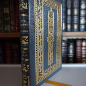 May include: A dark blue hardcover book with gold accents and the title "Lincoln Unbound" by Rich Lowry. The book is part of a signed limited edition series.
