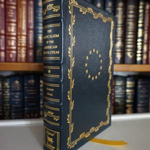 May include: A dark blue hardback book with gold lettering and a gold border. The title is "The Radicalism of the American Revolution" by Gordon Wood. The book is a signed edition.