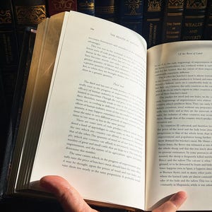 K&ouml;nnte beinhalten: Ein offenes Buch mit gealterten, vergilbten Seiten, gehalten von einer Hand. Der Text ist schwarz, mit einem dunkelblauen Einband. Der Hintergrund zeigt eine Sammlung antiker B&uuml;cher auf einem Regal.