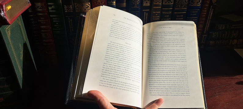 K&ouml;nnte beinhalten: Ein offenes Buch mit gealterten, vergilbten Seiten, gehalten von einer Hand. Der Text ist schwarz, mit einem dunkelblauen Einband. Der Hintergrund zeigt eine Sammlung antiker B&uuml;cher auf einem Regal.