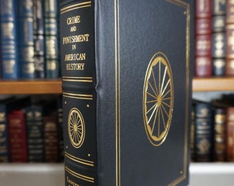 May include: A hardcover book titled "Crime and Punishment in American History" by Lawrence M. Friedman. The book has a dark blue cover with gold lettering and decorative accents, including a wheel-like emblem. The book is displayed on a shelf with other books.