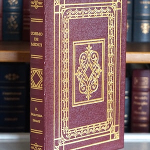 Puede incluir: Un libro de tapa dura burdeos titulado "Cosimo de Medici" de K. Dorothea Ewart. La portada presenta filigranas y letras doradas. El libro está de pie, con otros libros visibles en el fondo.