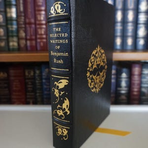 May include: A black hardcover book with gold lettering and decorative designs. The title reads "The Selected Writings of Benjamin Rush". The book is displayed on a shelf with other books in the background.