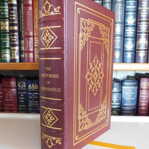 May include: A burgundy leather-bound book with gold accents and the title "The Histories of Herodotus" on the spine. The book is on a bookshelf with other books.
