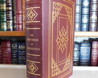 May include: A burgundy leather-bound book with gold accents and the title "The Histories of Herodotus" on the spine. The book is on a bookshelf with other books.