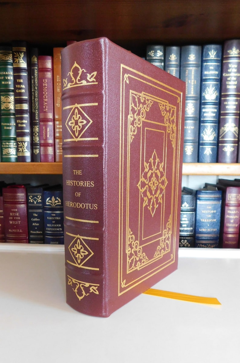May include: A burgundy leather-bound book with gold accents and the title "The Histories of Herodotus" on the spine. The book is on a bookshelf with other books.