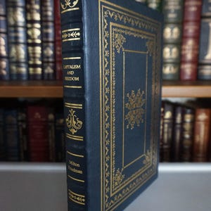 May include: A hardcover book titled "Capitalism and Freedom" by Milton Friedman. The book has a dark blue cover with gold decorative accents and lettering. The book is standing upright, showcasing its spine and front cover.