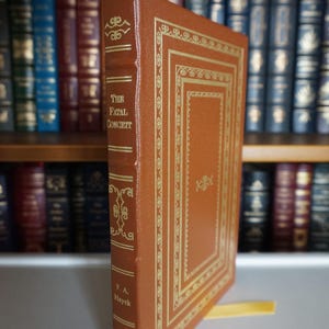 Puede incluir: Un libro de tapa dura marrón titulado "The Fatal Conceit" de F.A. Hayek. El libro tiene detalles decorativos y texto dorados. El libro está de pie, con otros libros visibles en el fondo.